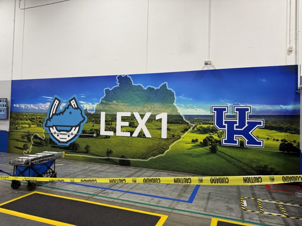 Wide side-angle view of Kentucky countryside mural with blue illustrated emblem, large LEX1 letters and UK logo inside warehouse.