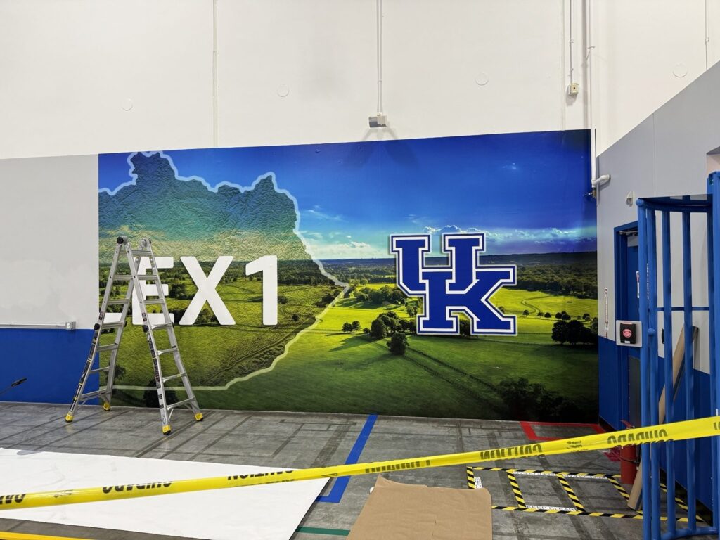 Warehouse wall with Kentucky landscape graphic, LEX1 lettering and UK emblem, with ladder and caution tape in front.