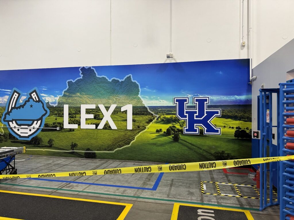 Front-facing view of an interior warehouse mural showing LEX1 text, Kentucky outline and large blue UK logo above caution tape.