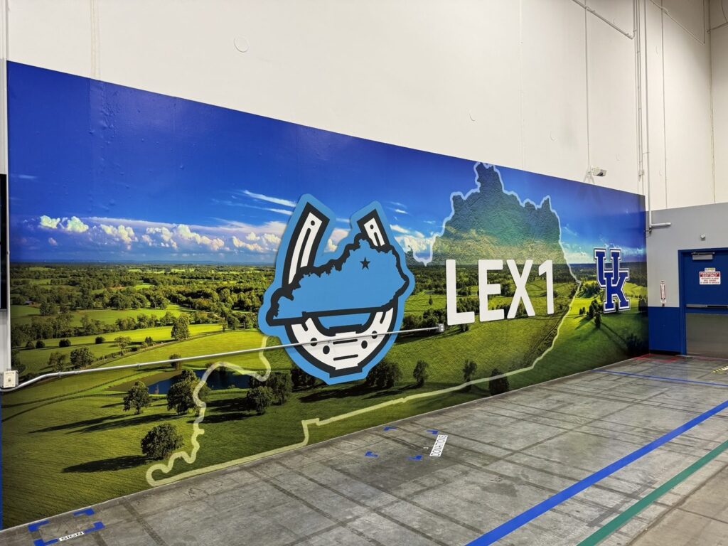 Angled view of expansive LEX1 warehouse wall graphic with Kentucky outline, green fields, blue illustrated emblem and University of Kentucky UK mark.