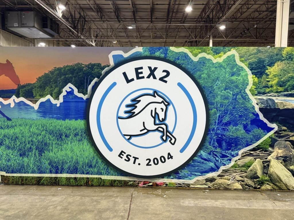 Close-up of circular LEX2 horse emblem with EST. 2004 lettering over green landscape and Kentucky outline on warehouse wall.