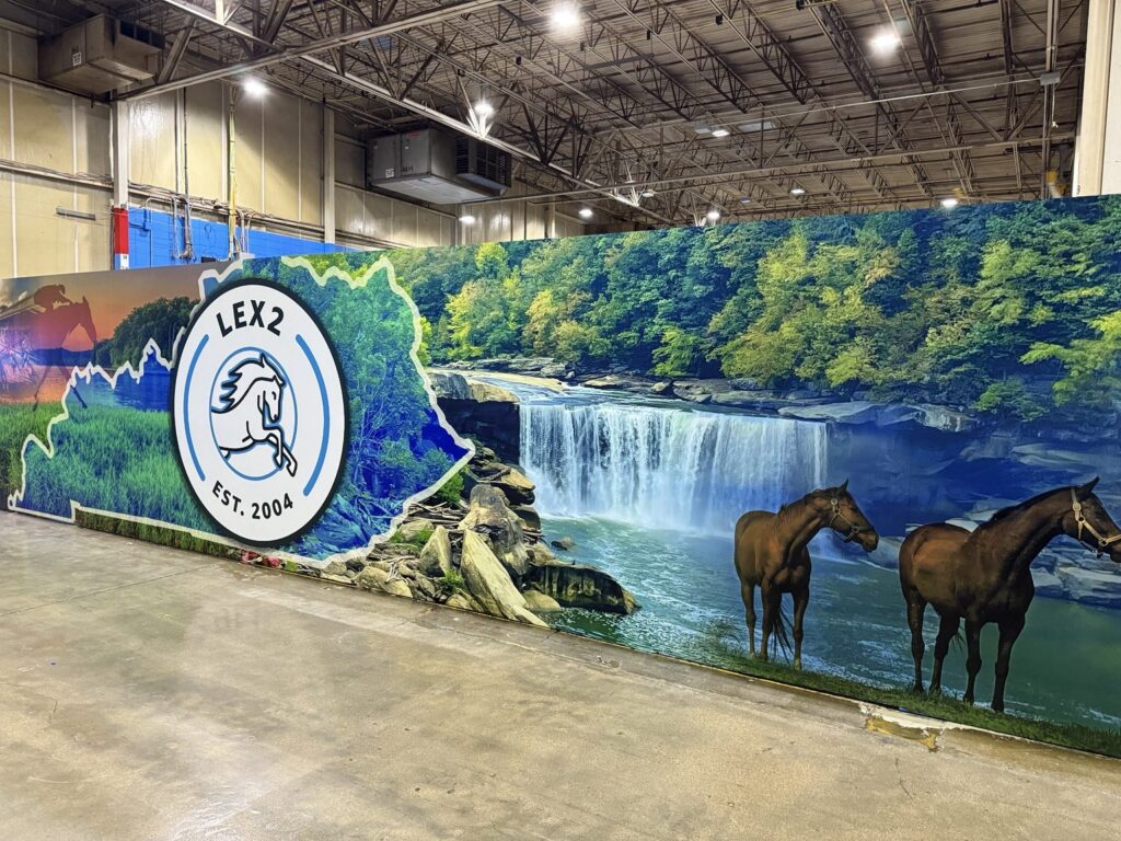 Long warehouse wall wrap showing wooded waterfall landscape and horses beside Kentucky outline and LEX2 logo.