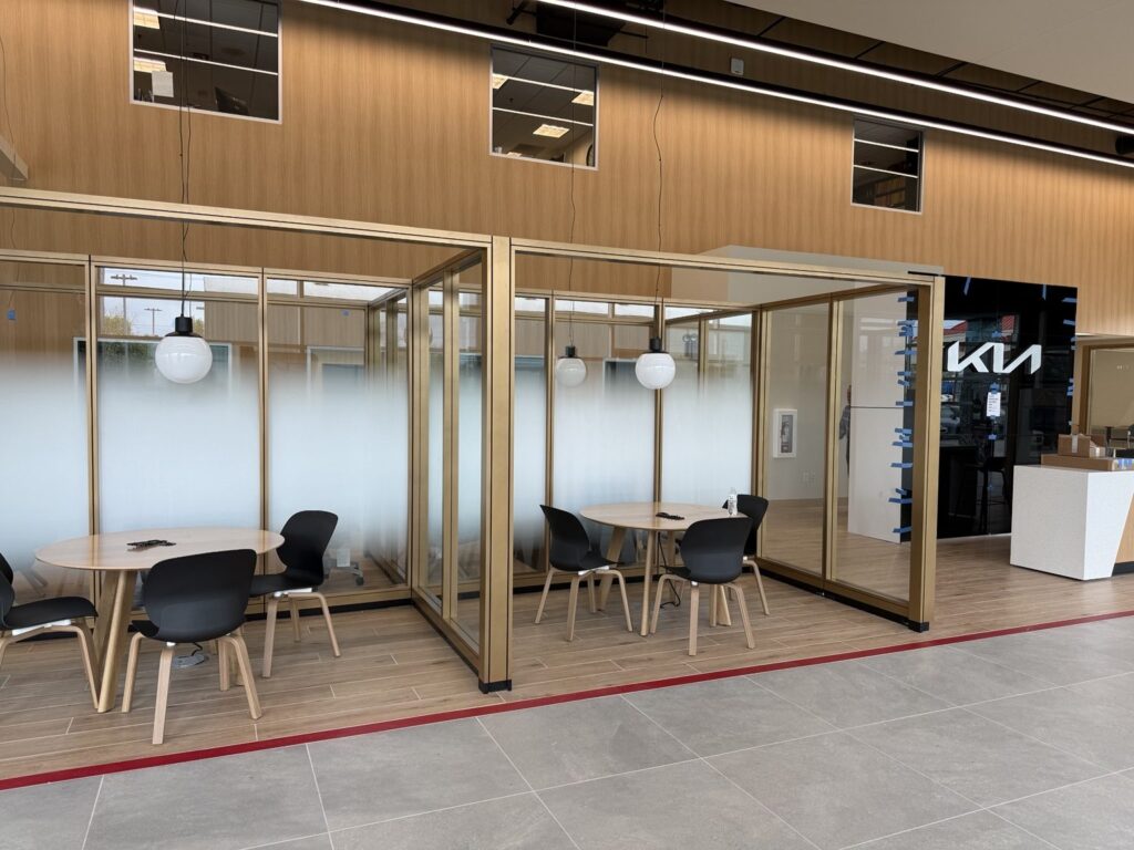 Interior of Kia dealership showing several glass meeting rooms with frosted gradient film, wood wall panels, black chairs and Kia logo in background.