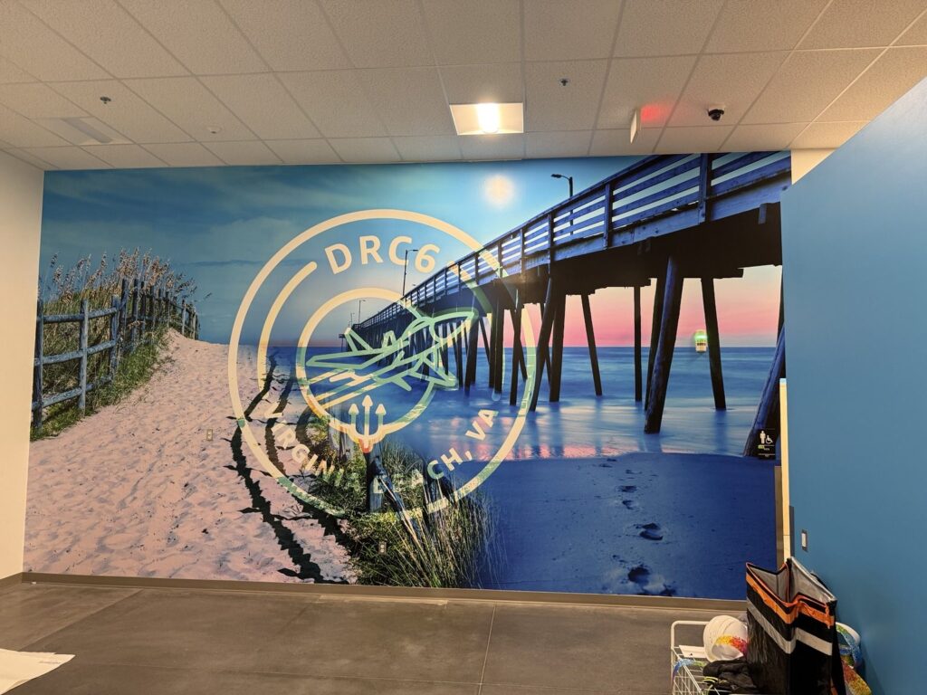 Full-wall coastal mural showing a beach path, ocean pier at sunset and DRC6 aviation insignia inside an Amazon facility.