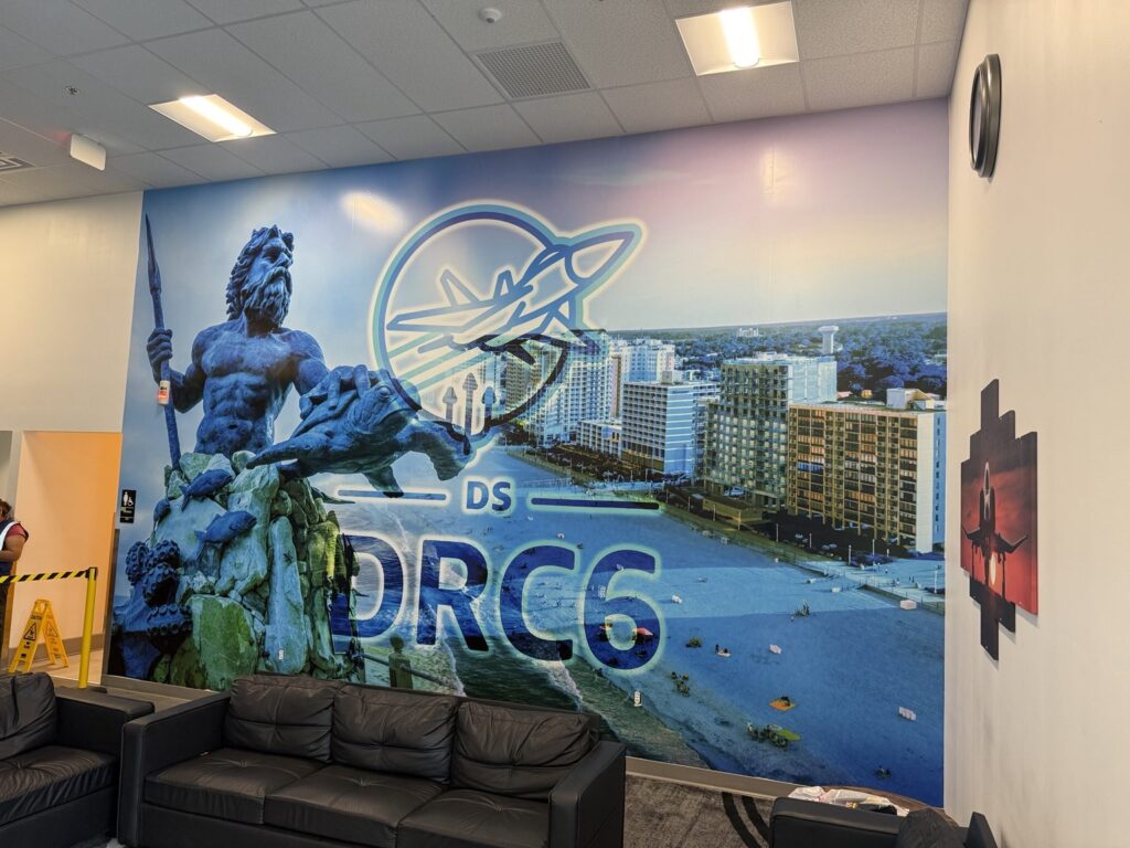 Interior wide view of Amazon DRC6 King Neptune coastal wall mural behind black lounge sofas with aircraft artwork on adjacent wall.