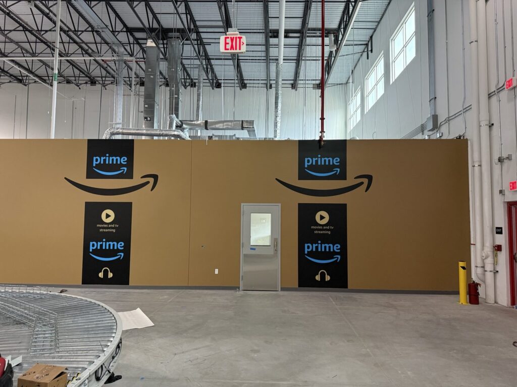 Straight-on view of tan shipping-facility wall featuring two large Amazon smile graphics and Prime panels positioned around a gray personnel door.