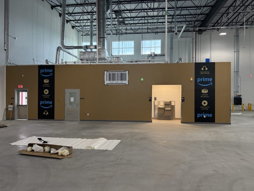 Ann Arbor wall graphics: wide view of Amazon Prime branded tan interior wall with black graphic panels, blue Prime logos, barcode, doors, and water-fountain alcove.