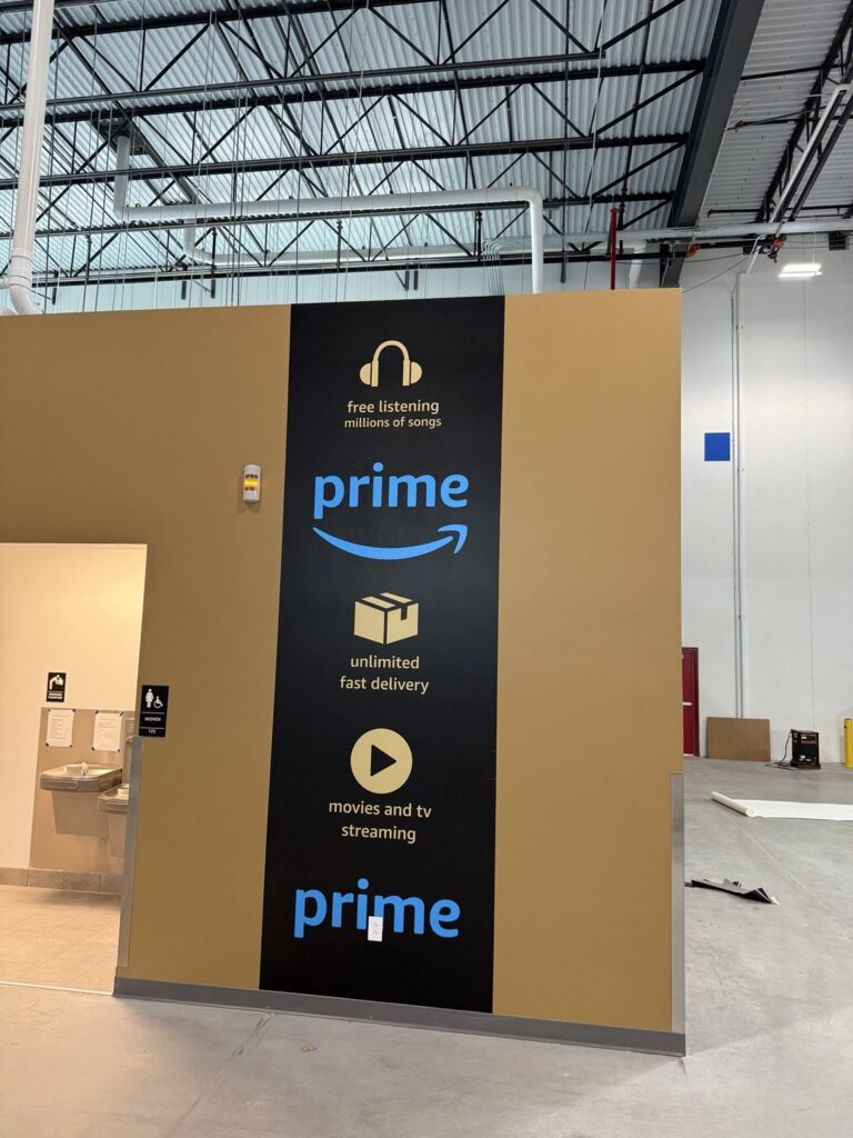 Tall black Prime wall graphic on tan industrial wall showing blue logos and icons for music, delivery, and video streaming.