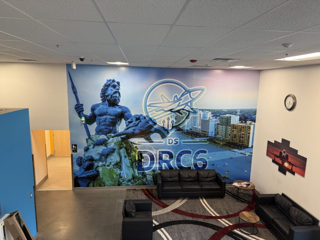 Virginia Beach wall wraps: wide view of large Amazon facility wall mural featuring King Neptune statue, Virginia Beach oceanfront, aviation emblem and DRC6 lettering above a seating area.
