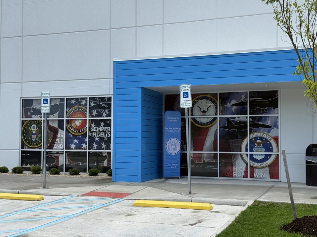 Exterior entrance view of Amazon facility with large military-themed window graphics featuring Army, Marine Corps, Navy and Air Force imagery.