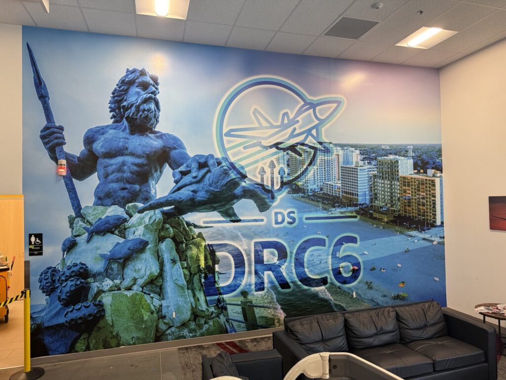Large interior mural of King Neptune, coastal buildings and DRC6 aviation logo behind black leather seating at an Amazon shipping facility.