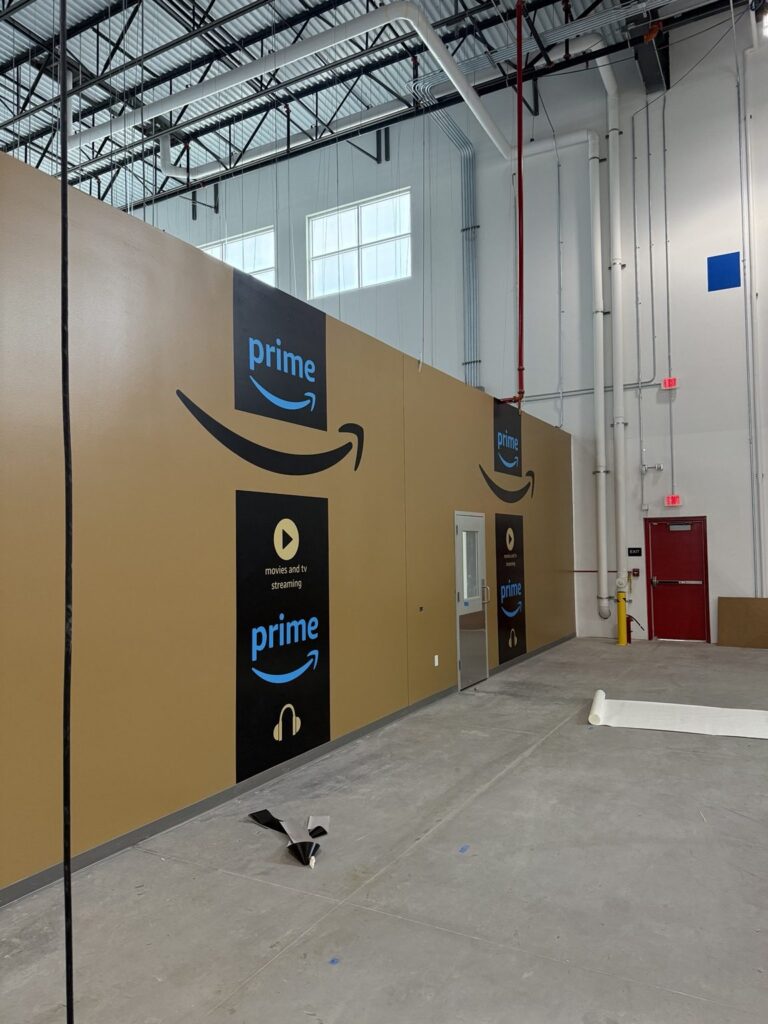 Angled view of an industrial interior wall with large Amazon smile logos and vertical black Prime branded graphic panels.