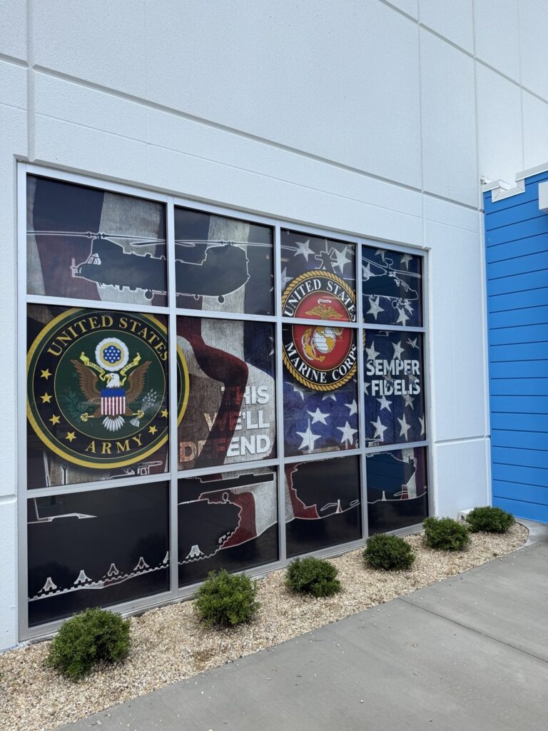 Angled exterior view of full-height window graphics displaying Army and Marine Corps emblems and helicopter silhouettes on an American flag-inspired background.
