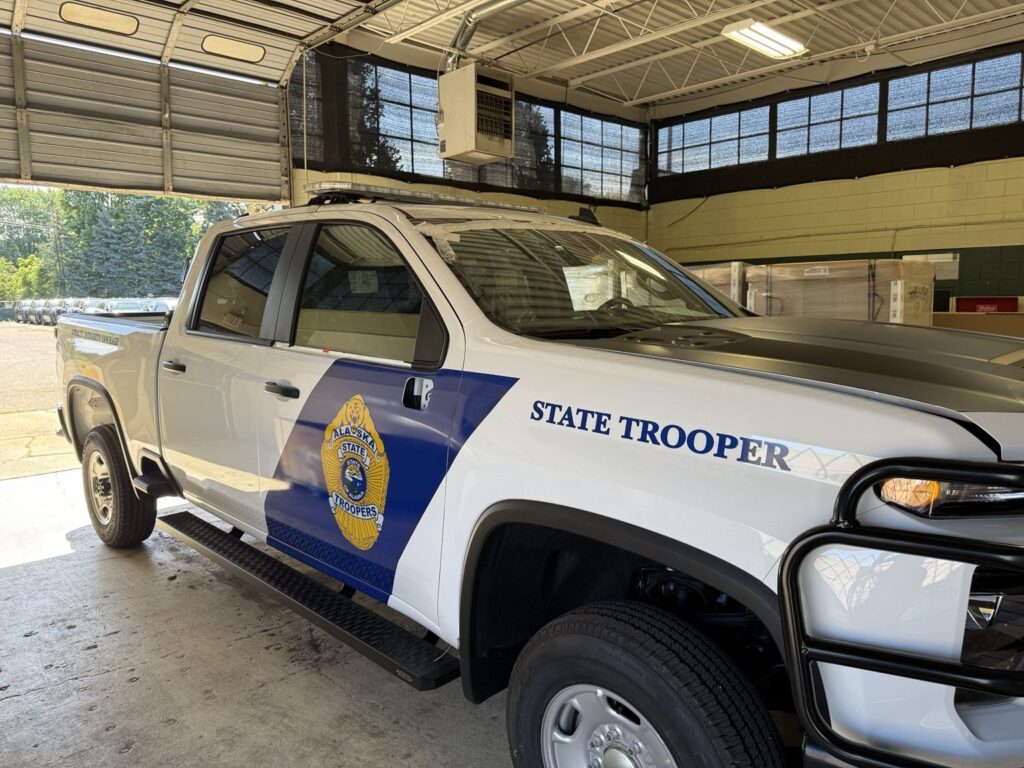 Three-quarter side view of Alaska State Troopers Chevrolet pickup with blue door graphics and gold badge inside a graphics shop
