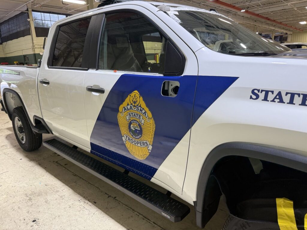 White Alaska State Troopers Chevrolet pickup during blue and gold door graphics installation, with door-handle opening visible