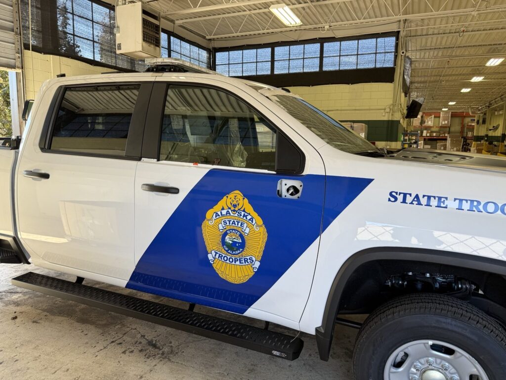 Side view of white Alaska State Troopers pickup in a vehicle graphics workshop with blue diagonal door stripe and gold emblem