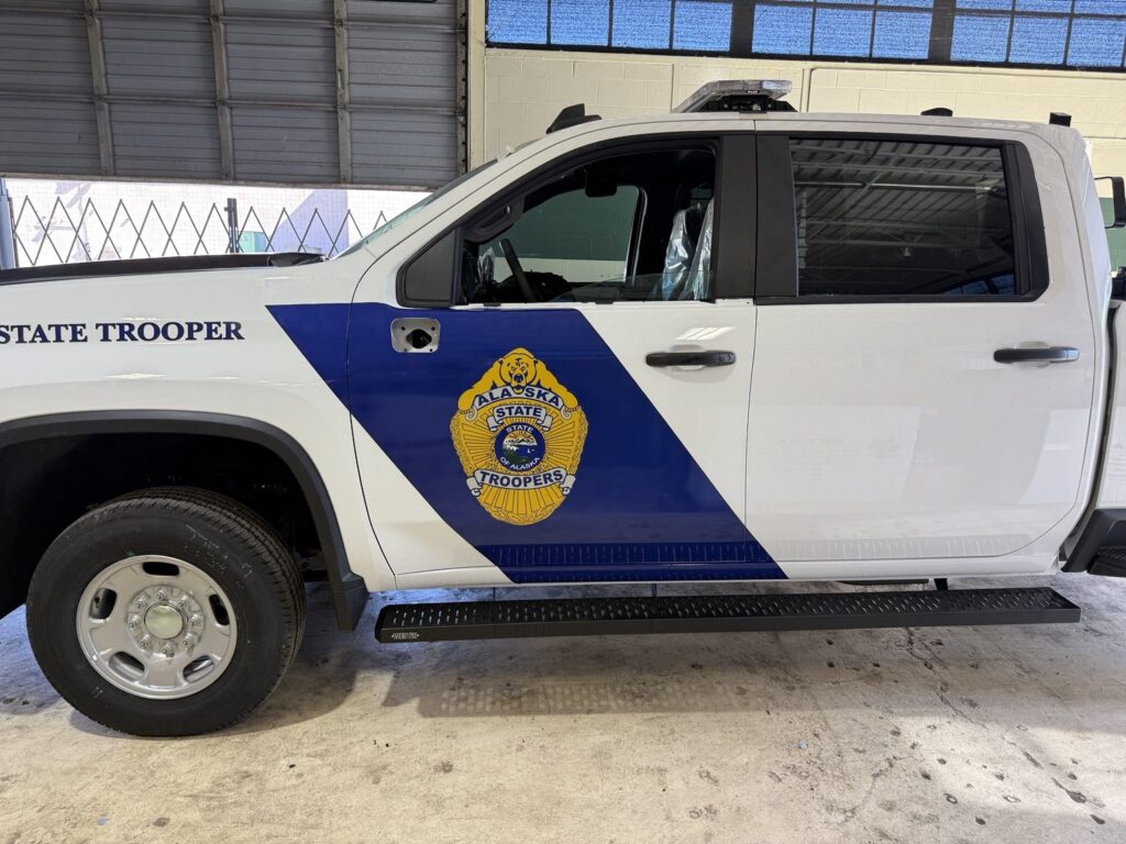Side profile of Alaska State Troopers white Chevrolet 2500HD with bold blue diagonal front-door graphic and gold badge