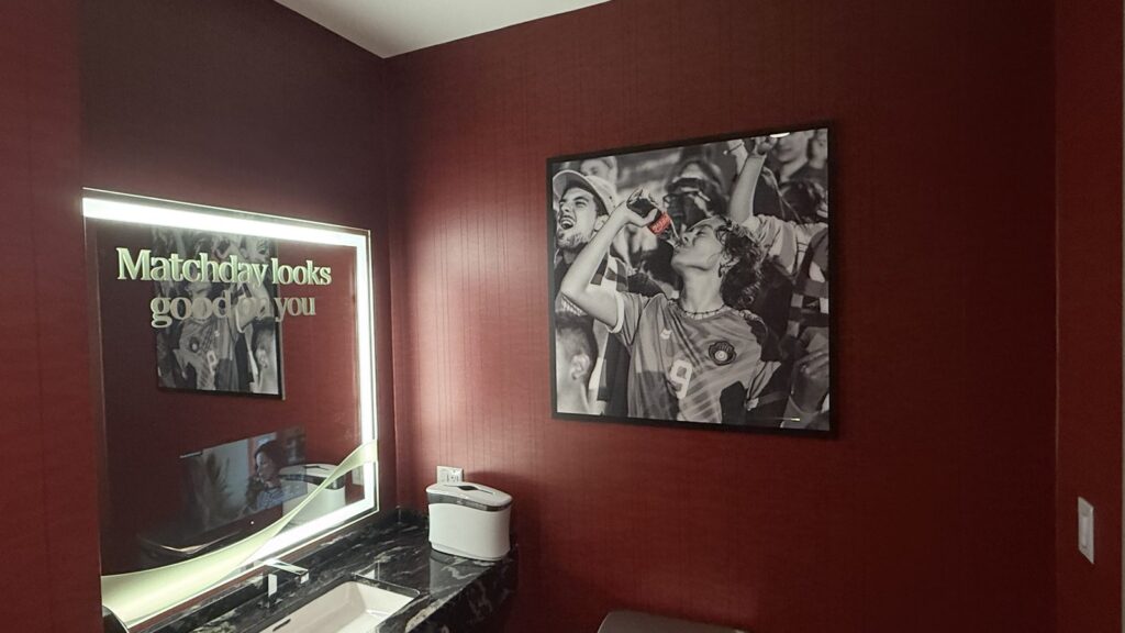 Mercedes-Benz Stadium suite restroom with a Matchday looks good on you mirror graphic and a framed Coca-Cola fan photo.