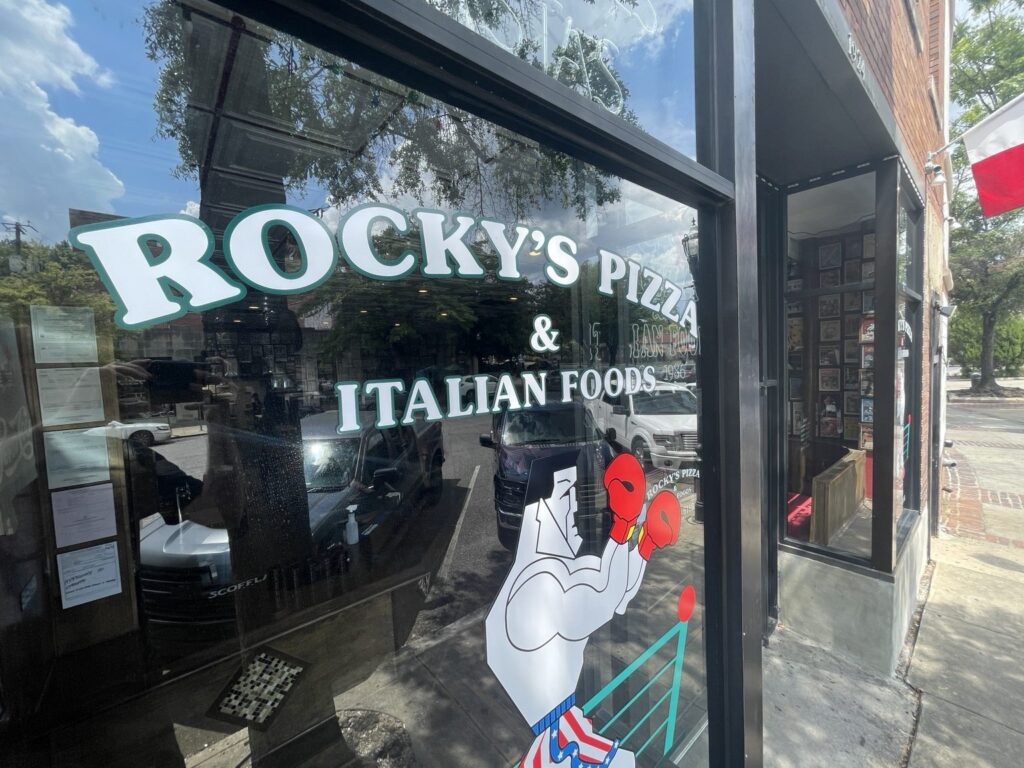 Angled view of Rocky’s Pizza & Italian Foods storefront window lettering and red-gloved boxer mascot in Five Points South, Birmingham.