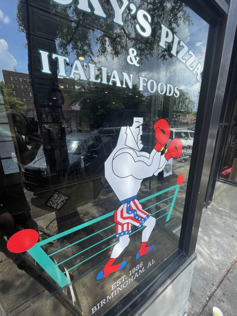 Close-up of Rocky’s Pizza boxing mascot with red gloves, patriotic shorts, turquoise boxing ring, and Birmingham Est. 1986 lettering on glass.