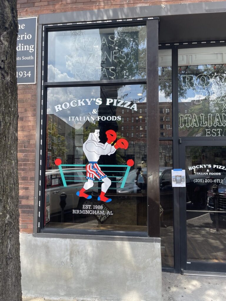 Left storefront window showing Rocky’s Pizza & Italian Foods white and green lettering above a cartoon boxer with red gloves.