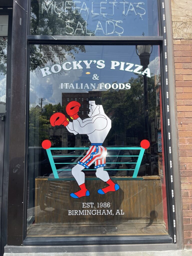 Front-facing view of right storefront window with Rocky’s Pizza & Italian Foods lettering, boxer illustration, turquoise ropes, and Birmingham Est. 1986 text.